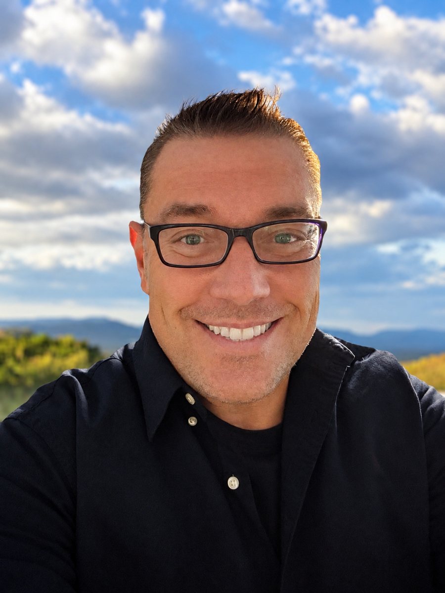 Man wearing black shirt and glasses smiling outdoors with mountains in background